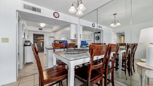Un coin repas avec une table en verre, des chaises en bois et des luminaires suspendus. On aperçoit une cuisine à l’arrière-plan, avec des meubles blancs et une horloge.