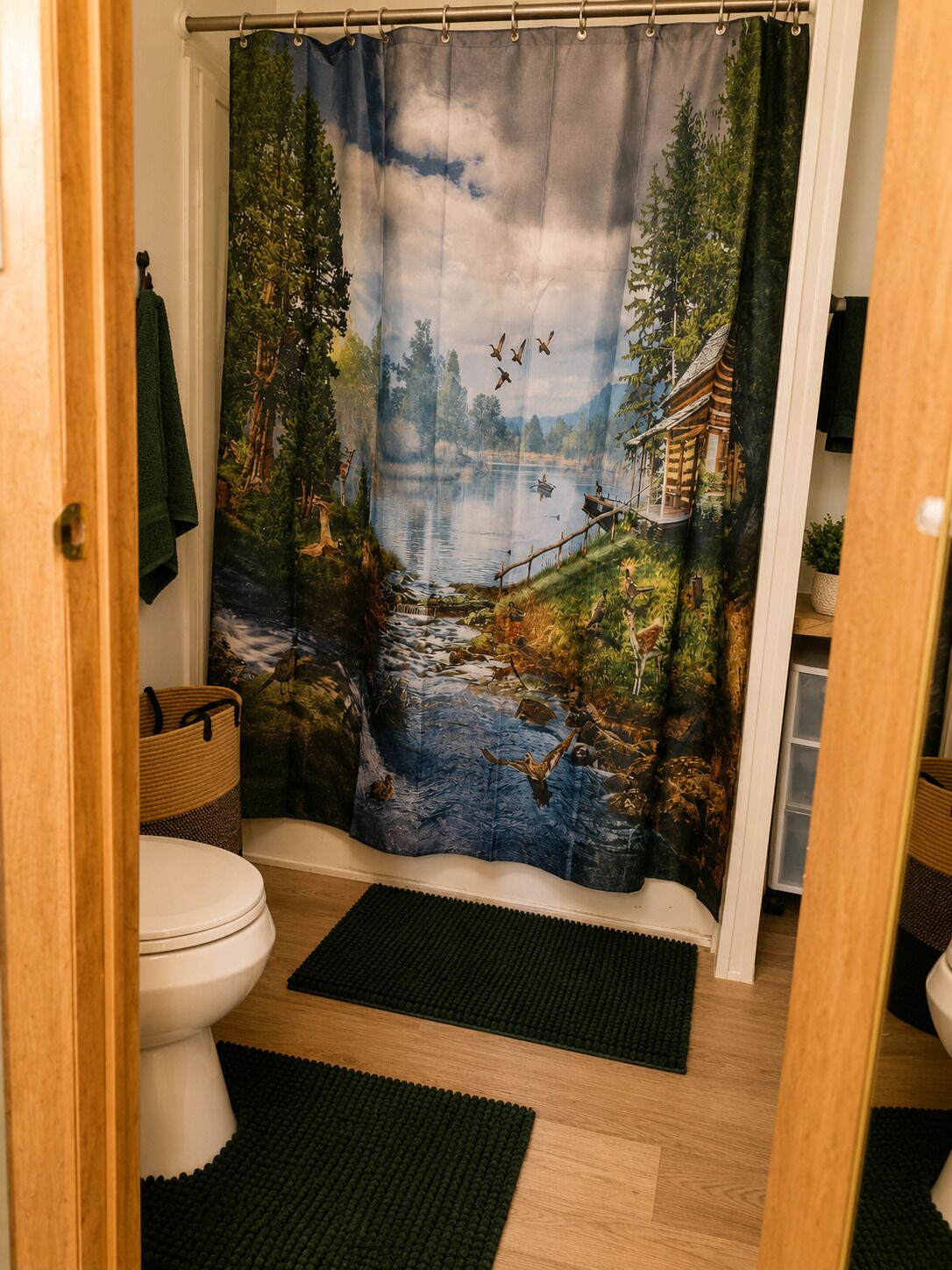 A bathroom with a shower curtain depicting a forest scene, a toilet, and two dark green mats on the floor.