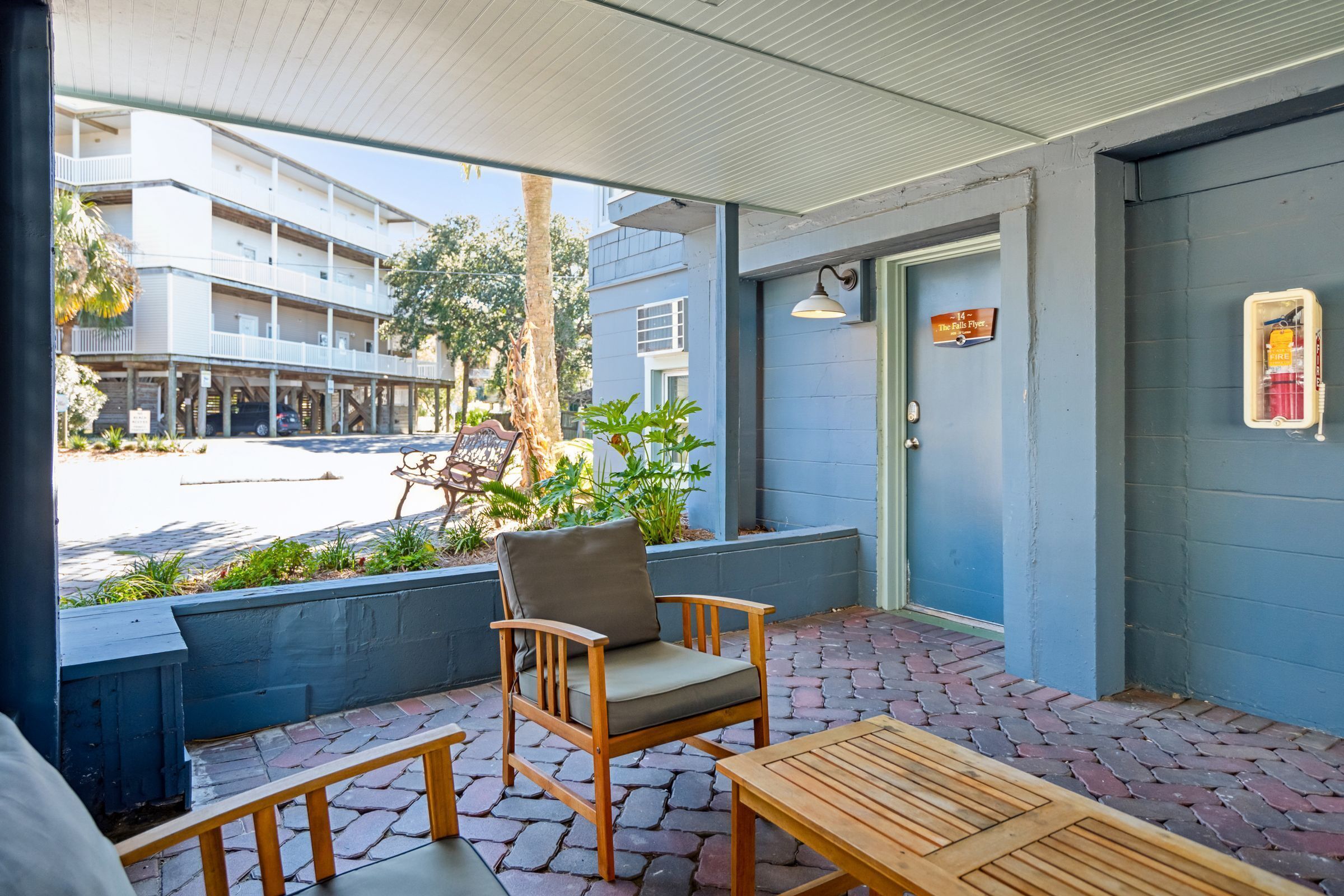 A covered outdoor seating area with a wooden chair and table, a blue building in the background, and a sign indicating the direction to the pool.
