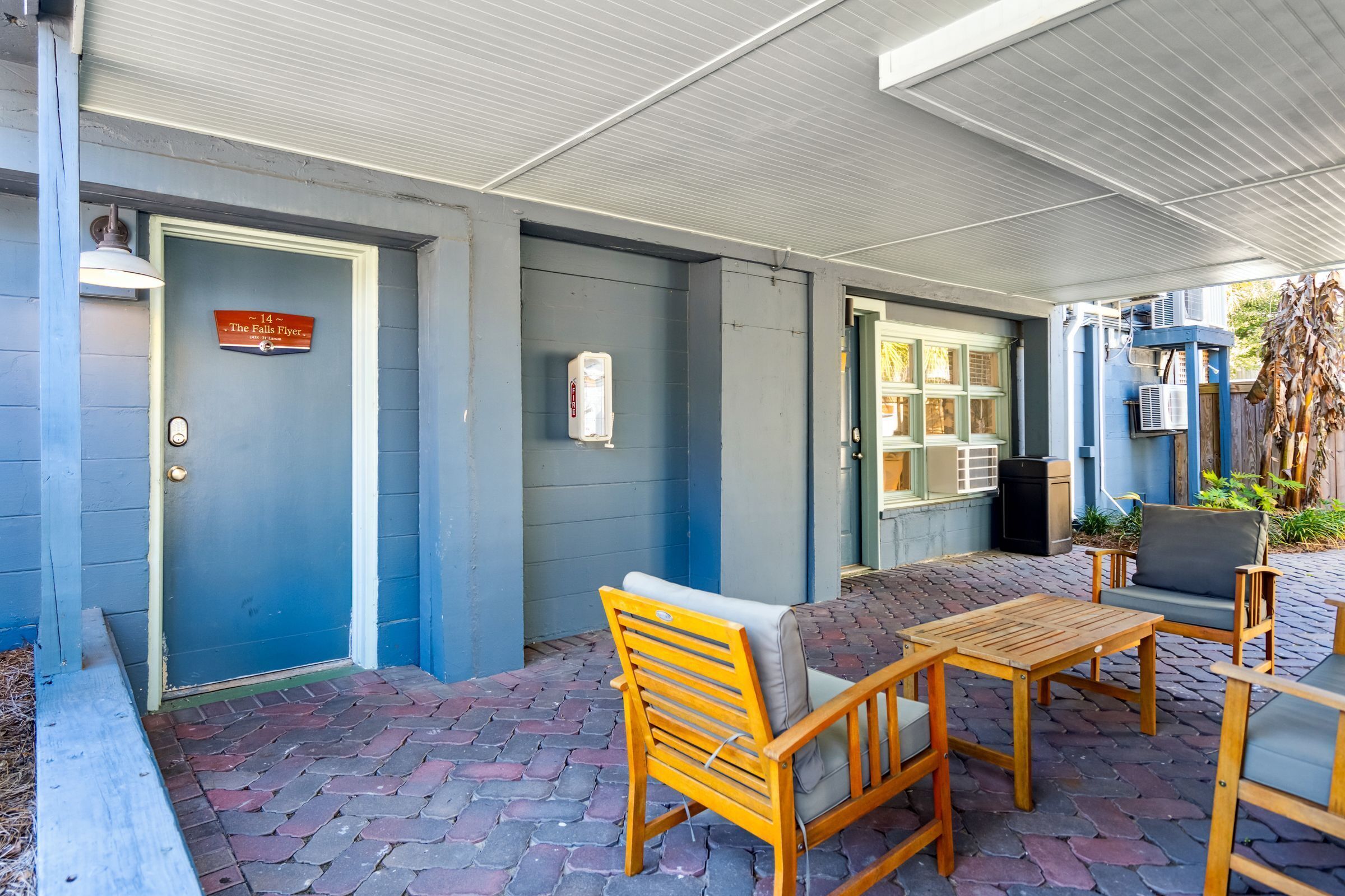 Un patio cubierto con mobiliario de madera, una puerta azul con un letrero y una ventana con estanterías.