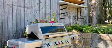Ein Edelstahlgrill mit vier Brennern und einem digitalen Display, vor einem Holzzaun, an dem ein Surfbrett befestigt ist.
