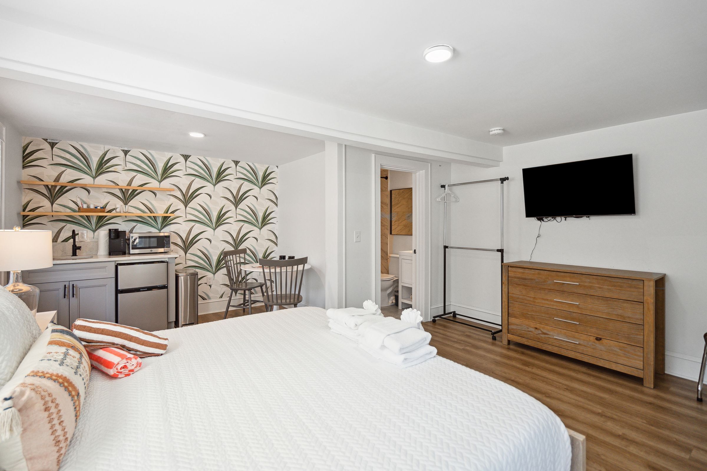 Une chambre d’hôtel avec un lit, un téléviseur fixé au mur, une commode en bois, un bureau avec une chaise et, au fond, une petite cuisine.