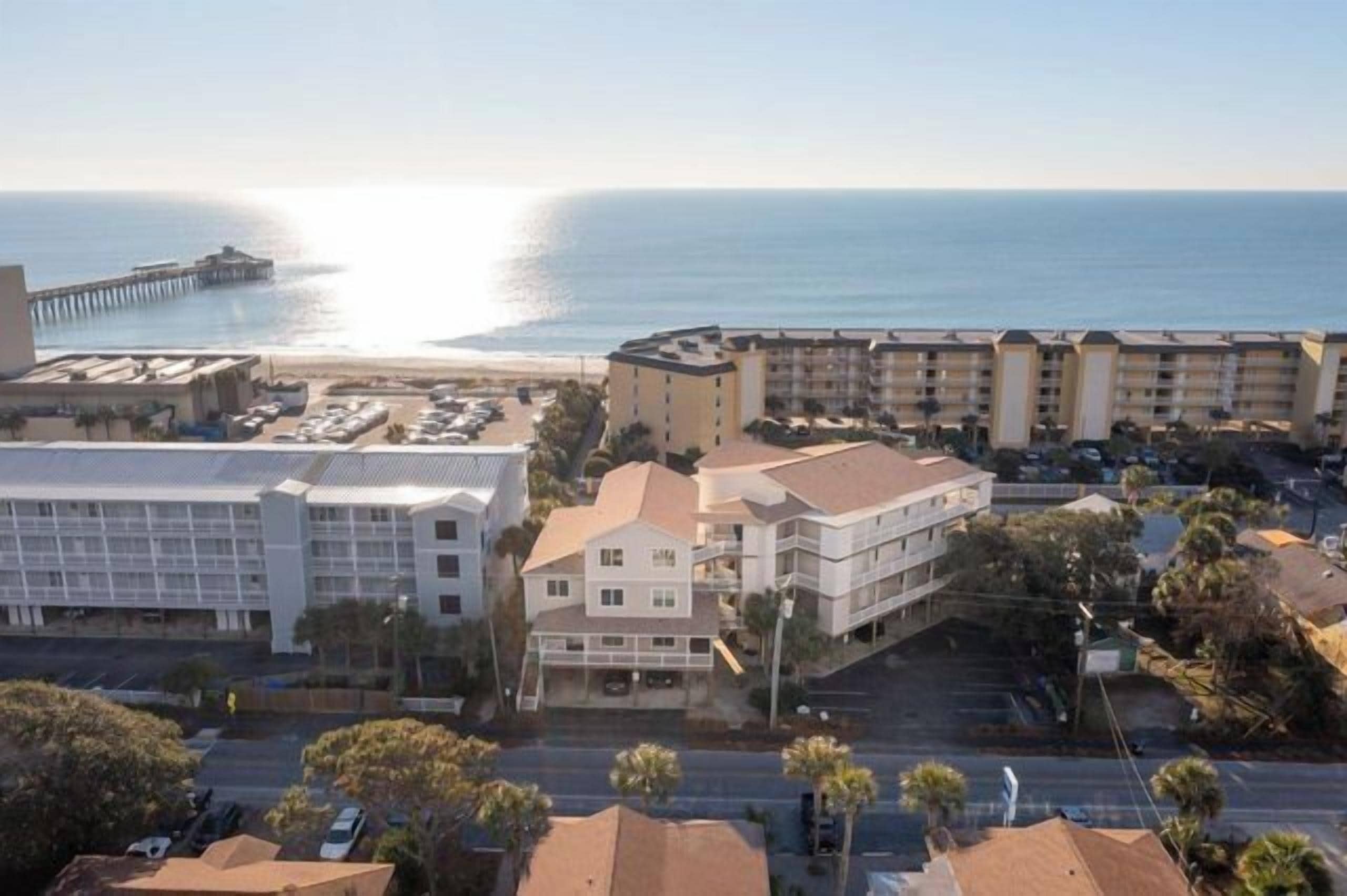 A coastal hotel with multiple buildings, a parking lot, and a pier in the background.