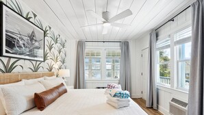 A neatly made bed with a quilted cover, a ceiling fan, and a large window with curtains. - Folliday Inn: The Torpedo: Room 22 (Folly Beach)