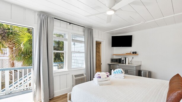 A bedroom with a bed, a TV, a desk, and a balcony with a view of palm trees. - Folliday Inn: The Torpedo: Room 22 (Folly Beach)