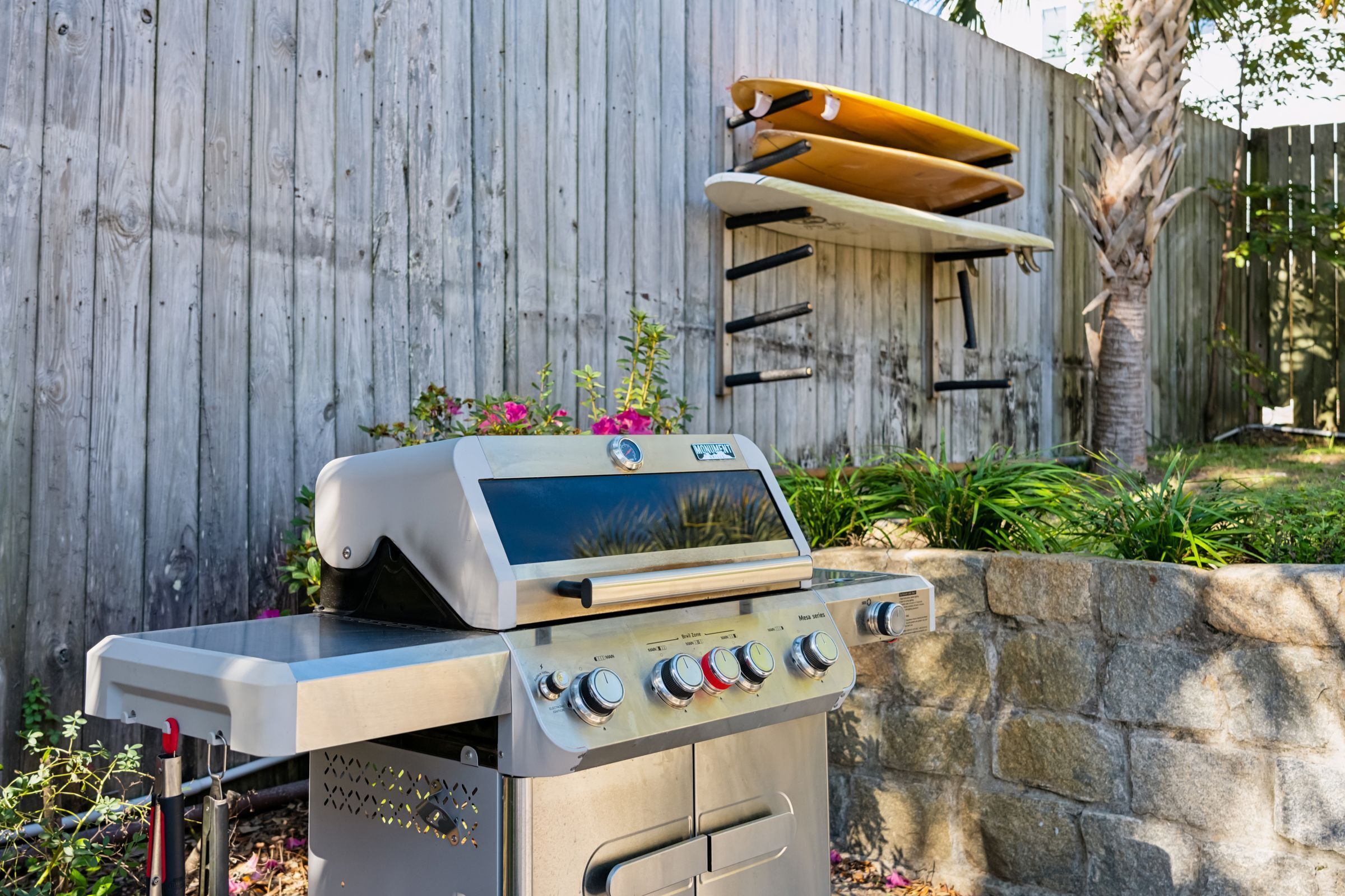 Una parrilla de acero inoxidable con cuatro quemadores y una pantalla digital, frente a una valla de madera con una tabla de surf colgada.