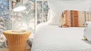A bedroom with a bed, white bedding, a woven side table, a wall-mounted lamp, and a tropical-themed wallpaper. - Folliday Inn - The Coronado - Room 11 (Folly Beach)