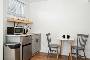 Een kleine keuken met een magnetron, fornuis en wastafel. Er staat een klein prullenbakje en een tafeltje met twee stoelen.