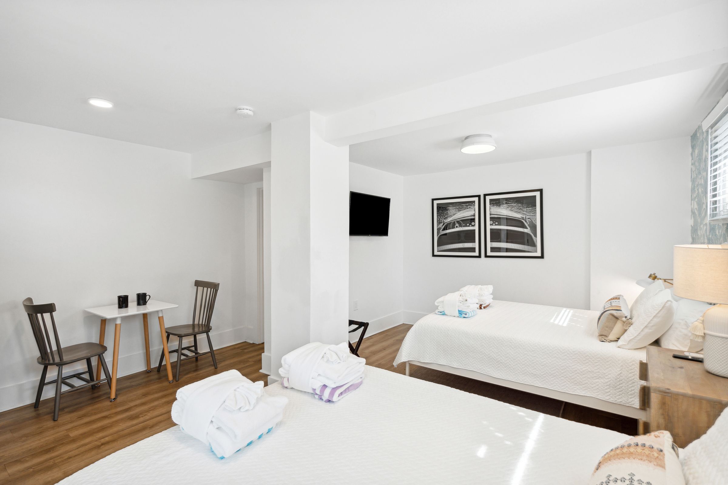 A hotel room with two beds, a small table with chairs, a TV mounted on the wall, and a window with blinds.