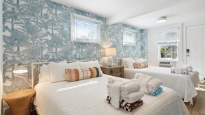 A hotel room with two beds, white bedding, a tropical wallpaper, and a small side table with a lamp.