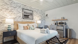 A bedroom with a bed, nightstands, a lamp, a picture on the wall, a microwave, and a small kitchen area. - Folliday Inn - The Triple - Room 24 (Folly Beach)
