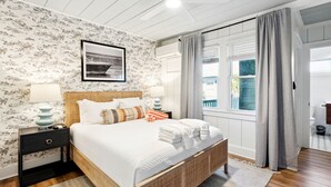 A bedroom with a large bed, a nightstand, a wall-mounted air conditioner, and a window with curtains. - Folliday Inn- The Overnighter- Room 25 (Folly Beach)