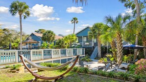 A hammock in a garden area with a pool and lounge chairs, surrounded by palm trees and a blue building. - Folliday Inn- The Overnighter- Room 25 (Folly Beach)