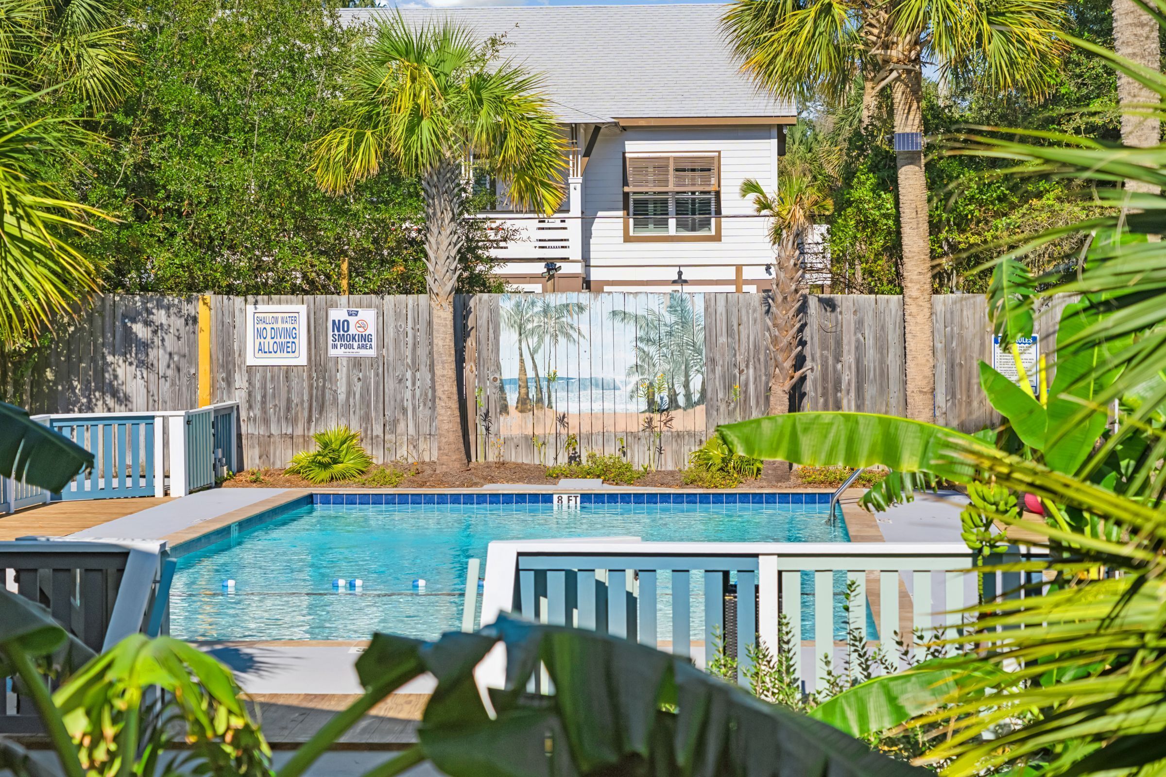 A swimming pool surrounded by a wooden fence, palm trees, and a house with a white exterior.