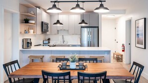 A modern kitchen with white cabinets, stainless steel appliances, and a wooden dining table with chairs. There are pendant lights above the table and a small bench in the corner. - Farwell's Spike by Revelstoke Vacations (Revelstoke)