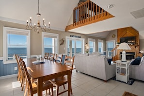 A spacious living area with a dining table, chairs, a sofa, and a TV. There are large windows offering a view of the sea.