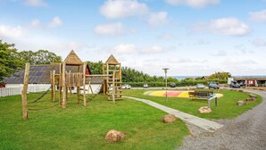 A playground with wooden climbing structures and a slide, surrounded by a grassy area with a paved path and a bench. - Awesome apartment in Allinge with WiFi (Allinge)
