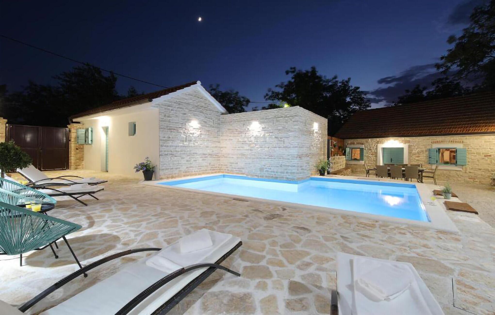 A stone pool with lounge chairs, a stone wall, and a small building with a tiled roof.