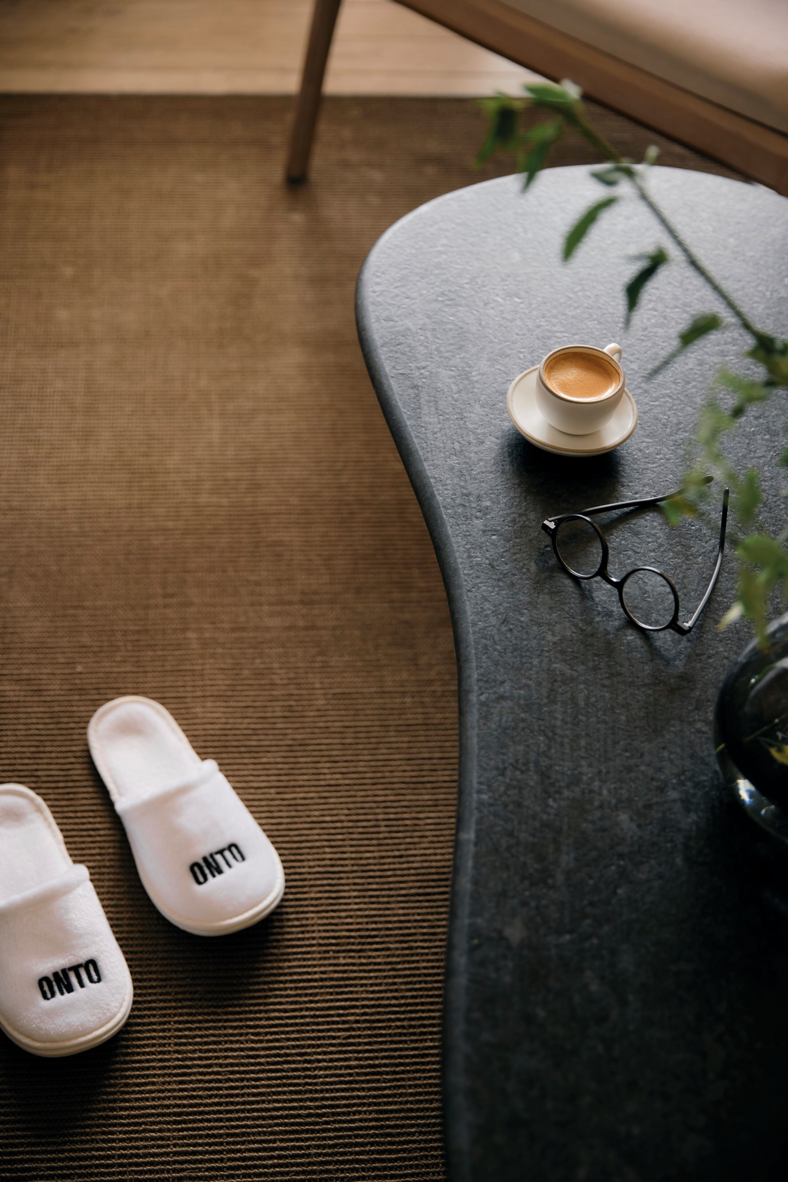 A pair of white slippers with 'ONTO' text, a cup of coffee, a pair of glasses, and a plant on a textured surface.