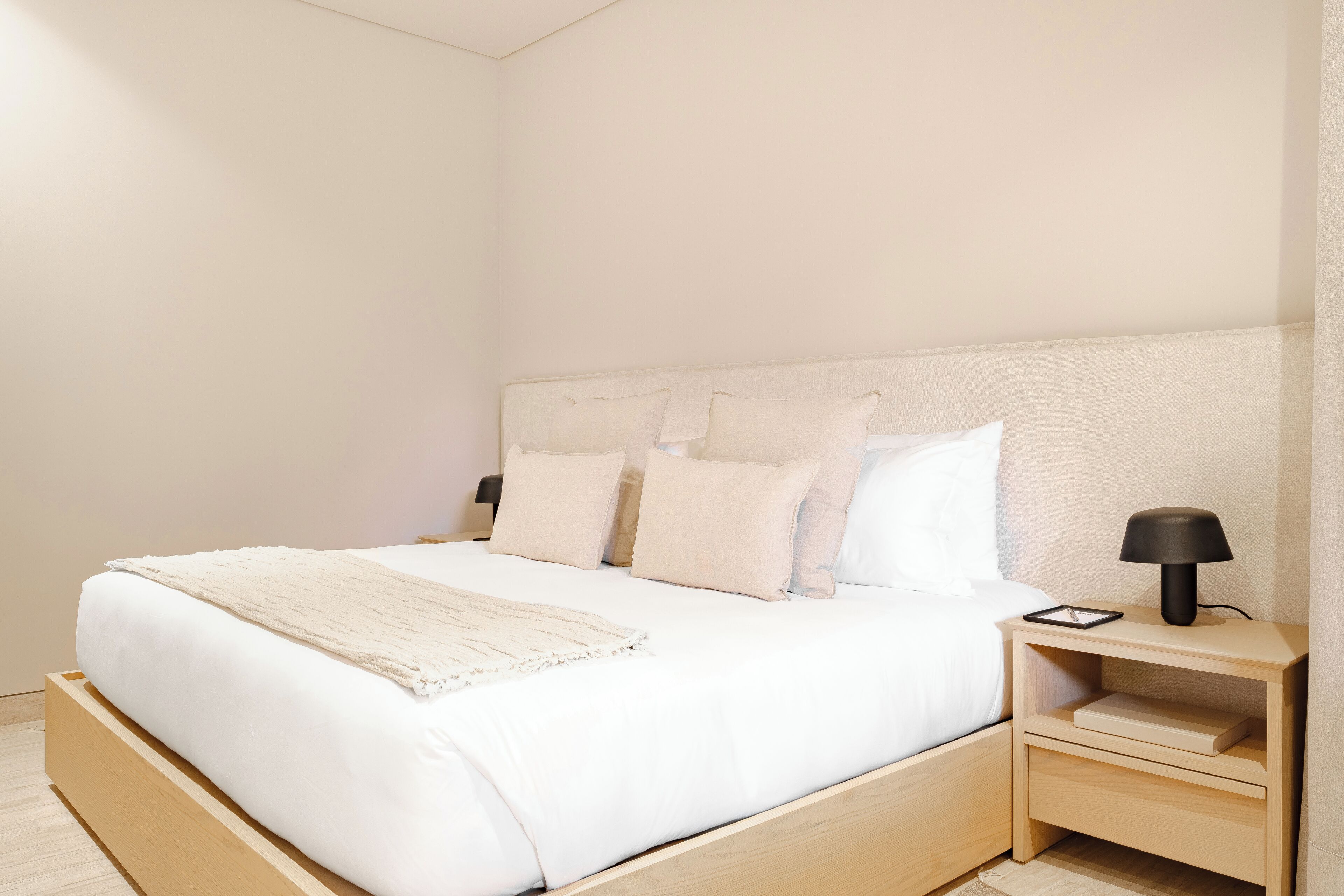 A minimalist bedroom with a wooden bed, white bedding, and a bedside table with a lamp.