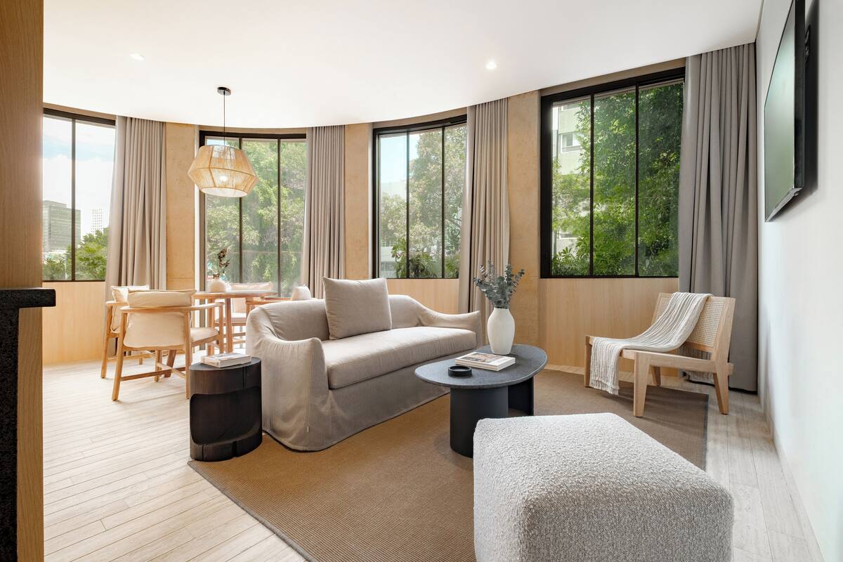Una sala de estar moderna con sofá, sillón y mesa de centro, ventanas grandes con cortinas, y una zona de comedor con muebles de madera.