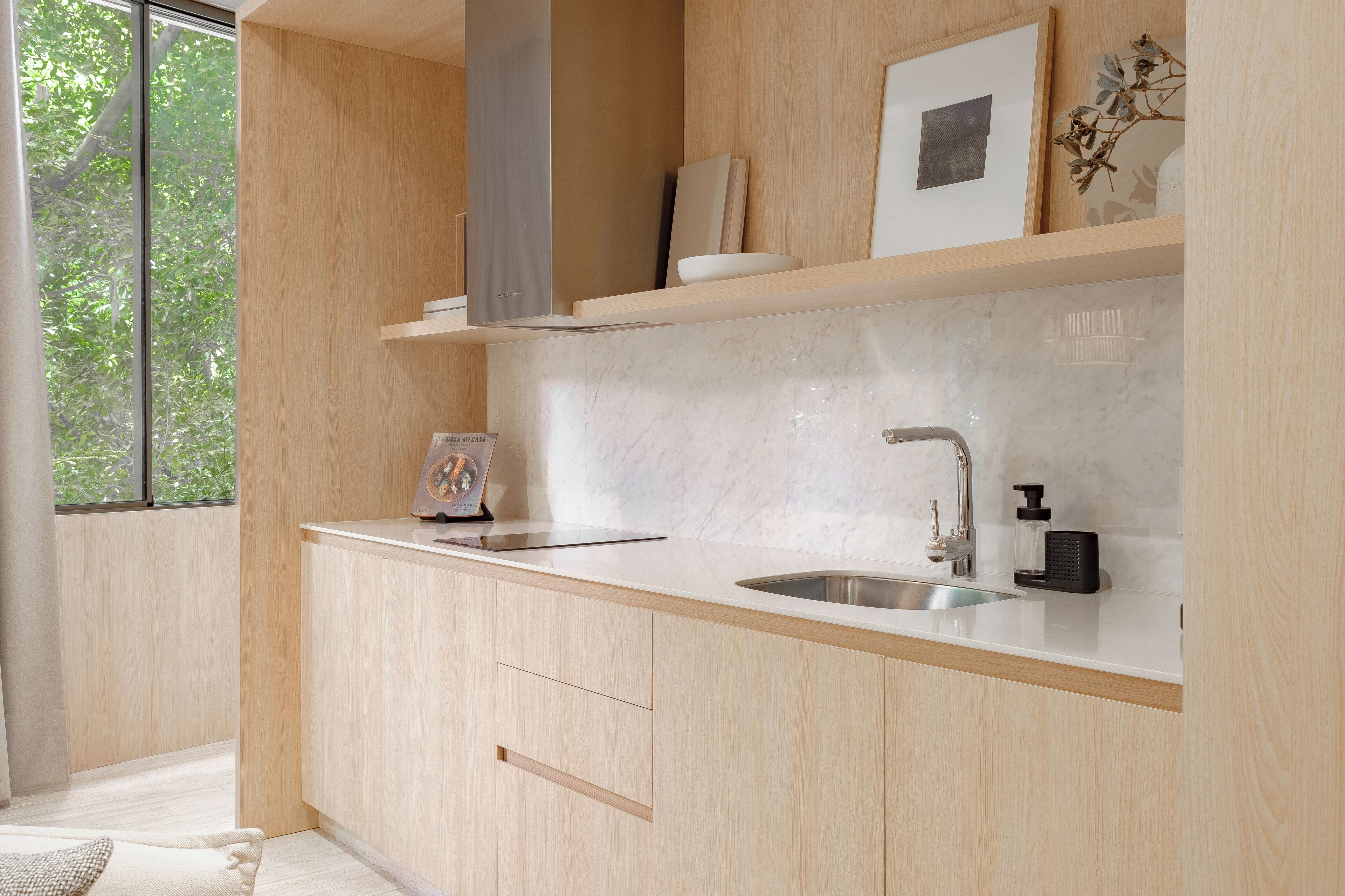 A modern kitchen with light wood cabinets, a white countertop, a stainless steel sink, and a window providing a view of greenery outside.