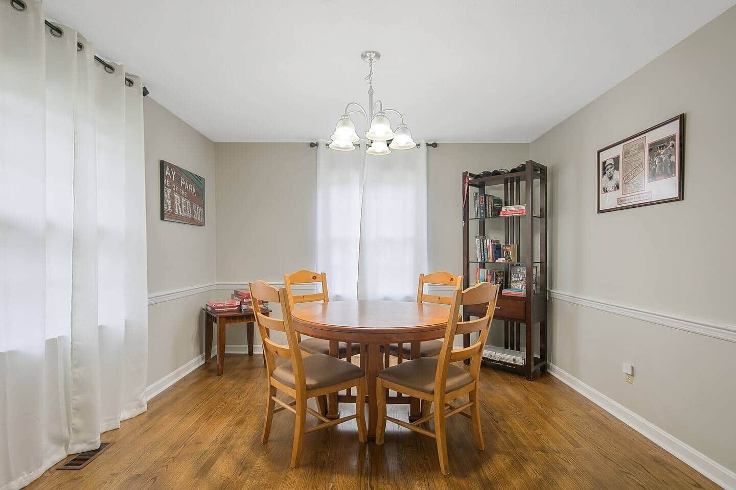 Un comedor con una mesa y sillas de madera, una araña, una estantería y cuadros en las paredes.