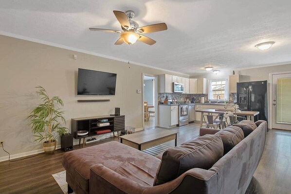 A modern living room with a flat-screen TV, a ceiling fan, a sofa, a dining area, and a kitchen in the background. - Home Base (Fayetteville)