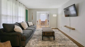 Un salon avec un canapé, une table basse, un téléviseur fixé au mur et, au fond, un coin repas.