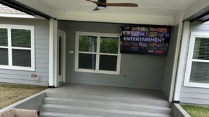 Een overdekt terras met een plafondventilator, een flatscreen televisie aan de wand bevestigd en beige zachte zitplaatsen.