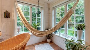 Una habitación con hamaca, silla de mimbre, plantas en macetas y ventanas amplias con vistas a la vegetación.