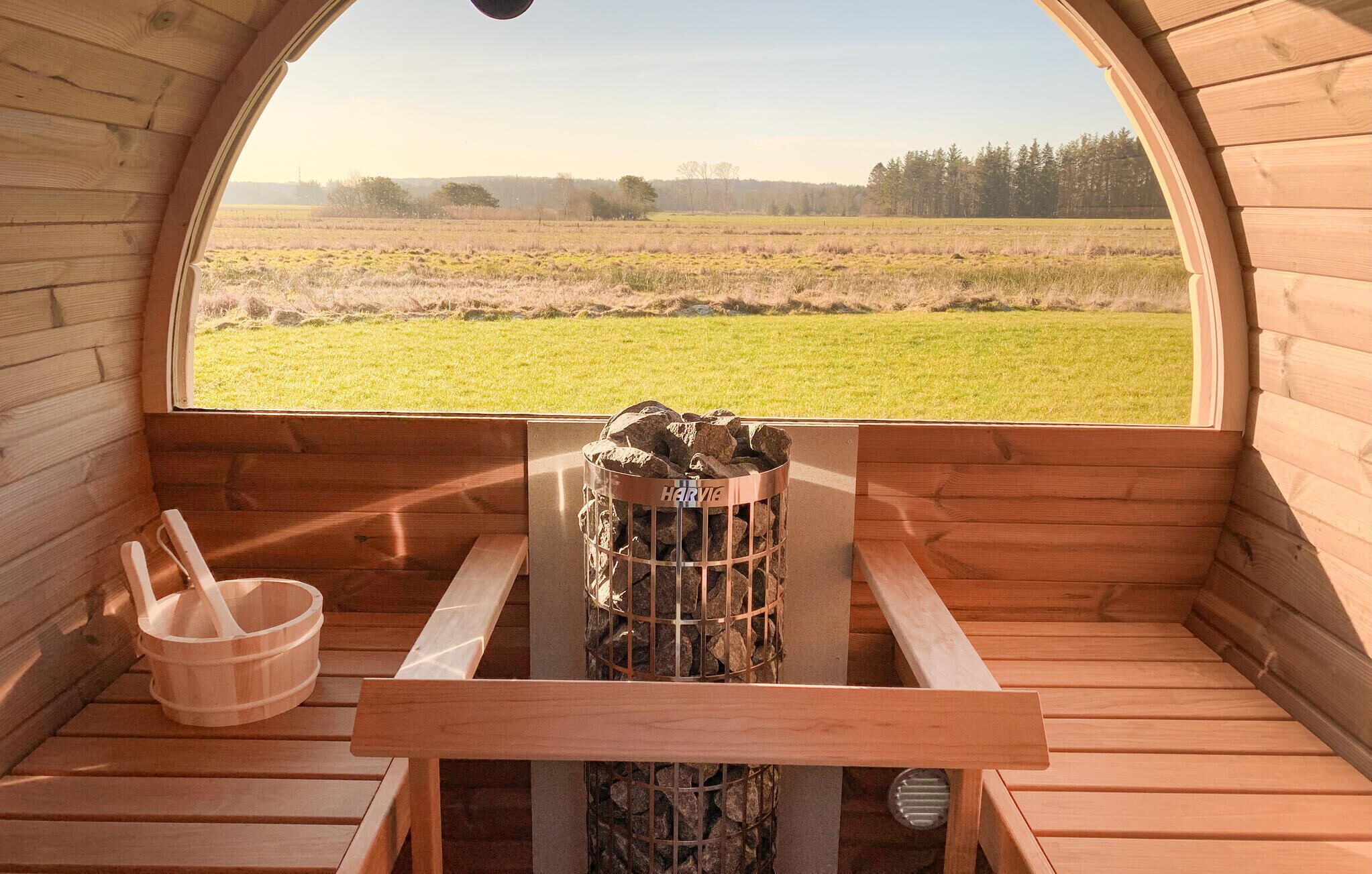 Una cabana de fusta amb una panxa i un contenidor metàl·lic amb l'etiqueta “HARVIG” a terra, un cubell de fusta, i vista a un camp herbós a través d'una finestra gran i arquejada.