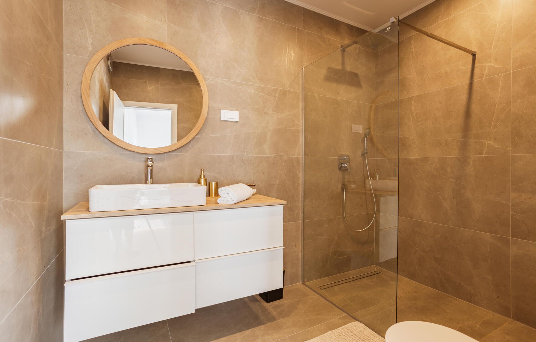 Un baño moderno con ducha envuelta en vidrio, un espejo redondo y un tocador blanco con cajones.