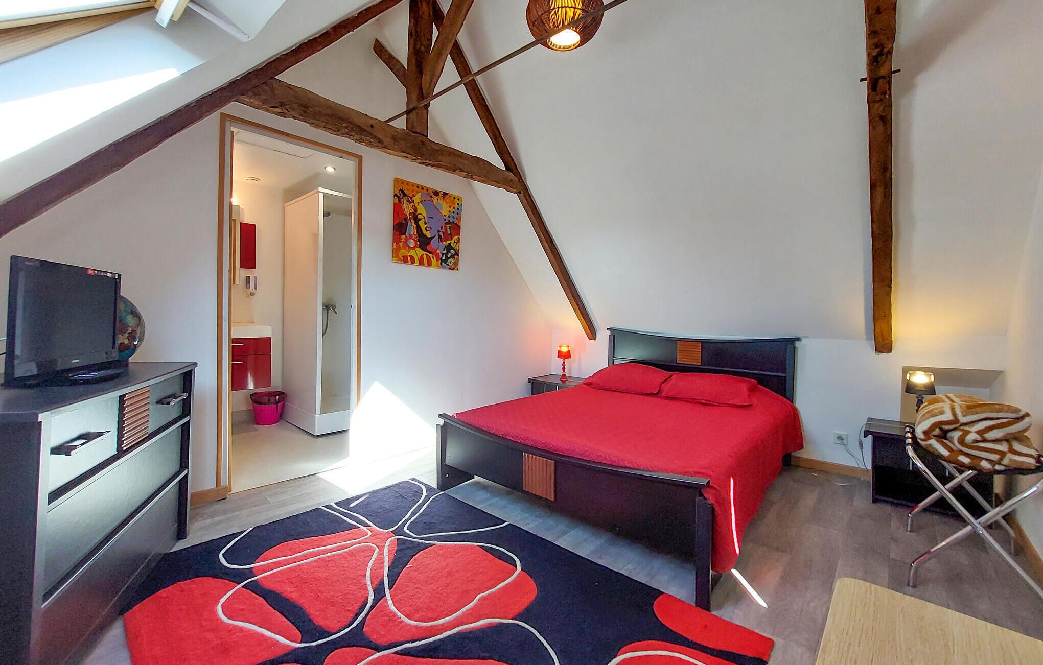 A bedroom with a sloped ceiling, a bed with red bedding, a desk with a computer, a chair, and a small table with a lamp.
