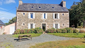 Une maison en pierre avec une allée gravillonnée, un banc en bois et un parterre de fleurs devant.