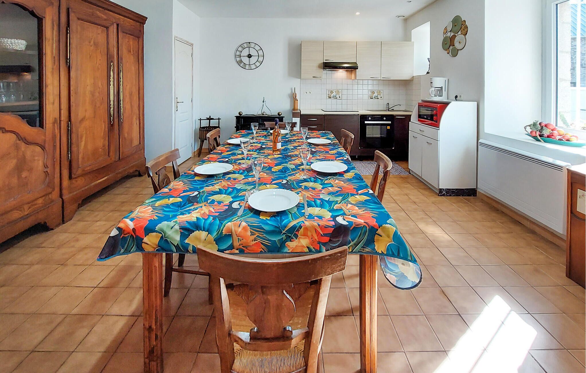 A dining area with a long table set for six, wooden chairs, a wooden cabinet, a kitchen in the background, and a clock on the wall.