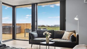 A modern living room with a black leather sofa, a small round table, and a vase of flowers. Large glass doors lead to an outdoor patio with a wooden deck and a view of the countryside. - Amazing home in Børkop with sauna (Børkop)