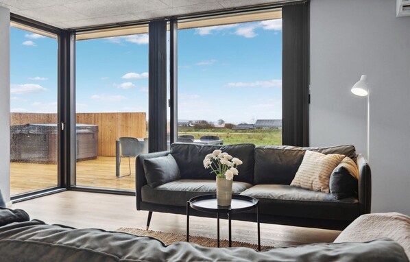 En moderne stue med en sort lædersofa, et lille rundt bord og en vase med blomster. Store glasdøre fører ud til en udendørs terrasse med træterrasse og udsigt over landskabet.
