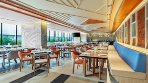 A spacious restaurant with multiple dining tables and chairs, a long counter with a blue bench, and large windows offering a view of greenery outside.