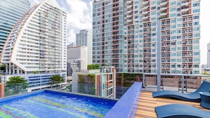 A rooftop pool area with a view of high-rise buildings, lounge chairs, and a wooden deck.