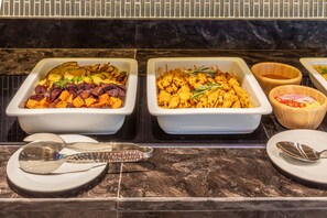 Buffet com diversos pratos, incluindo legumes assados e frango, sobre um balcão de mármore.