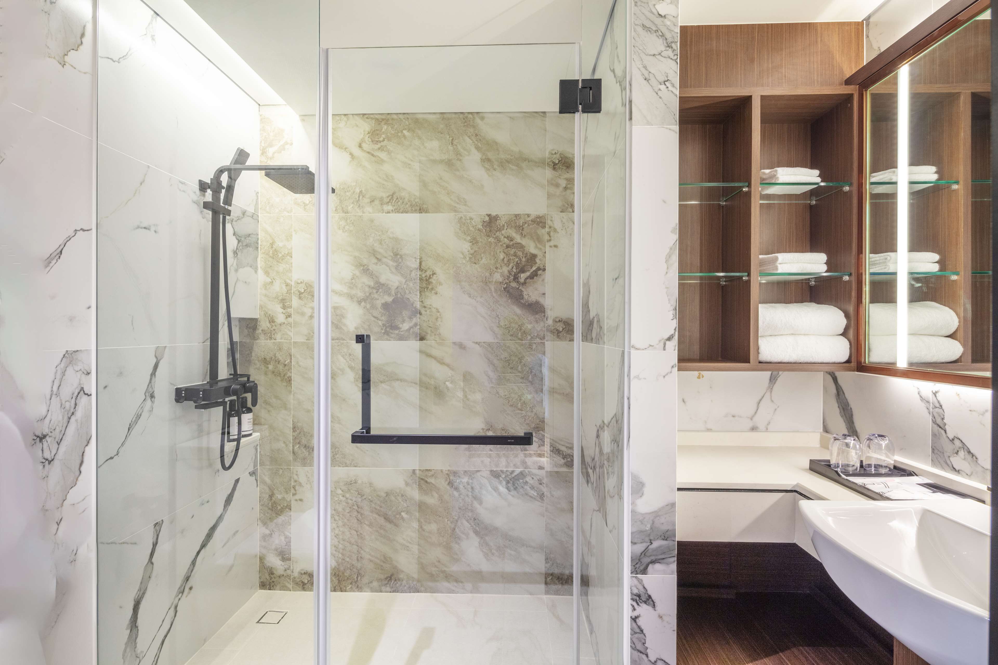 Un baño moderno con ducha de vidrio, paredes de mármol, un mueble de tocador de madera con repisas de vidrio y un lavamanos blanco.