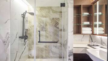 Un baño moderno con ducha de vidrio, paredes de mármol, un mueble de tocador de madera con repisas de vidrio y un lavamanos blanco.