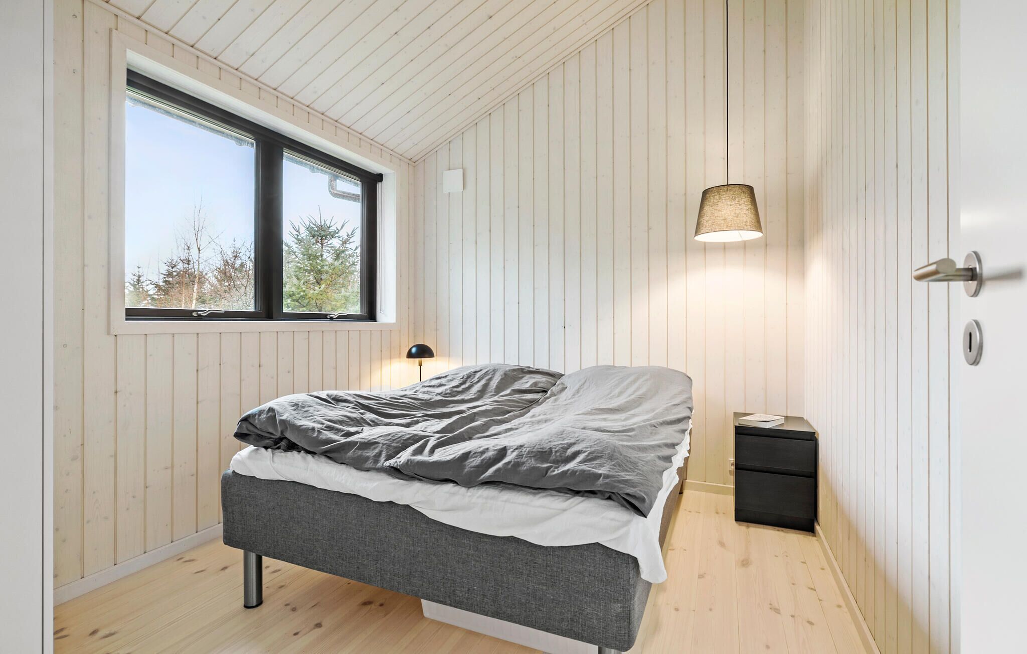 Ein Schlafzimmer mit einem Bett, einem Nachttisch, einer Lampe und einem Fenster mit Blick auf Bäume.