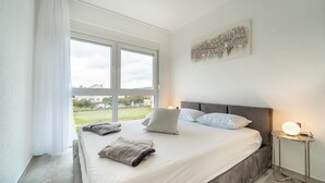 A bedroom with a large window, a bed with pillows and a blanket, a bedside table with a lamp, and a wall art piece. - Beautiful apartment in Sukosan (Sukosan)