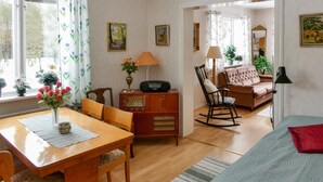 A cozy living room with a wooden table, chairs, a sofa, and a rocking chair. There is a radio on a wooden cabinet, a lamp, and a window with curtains.