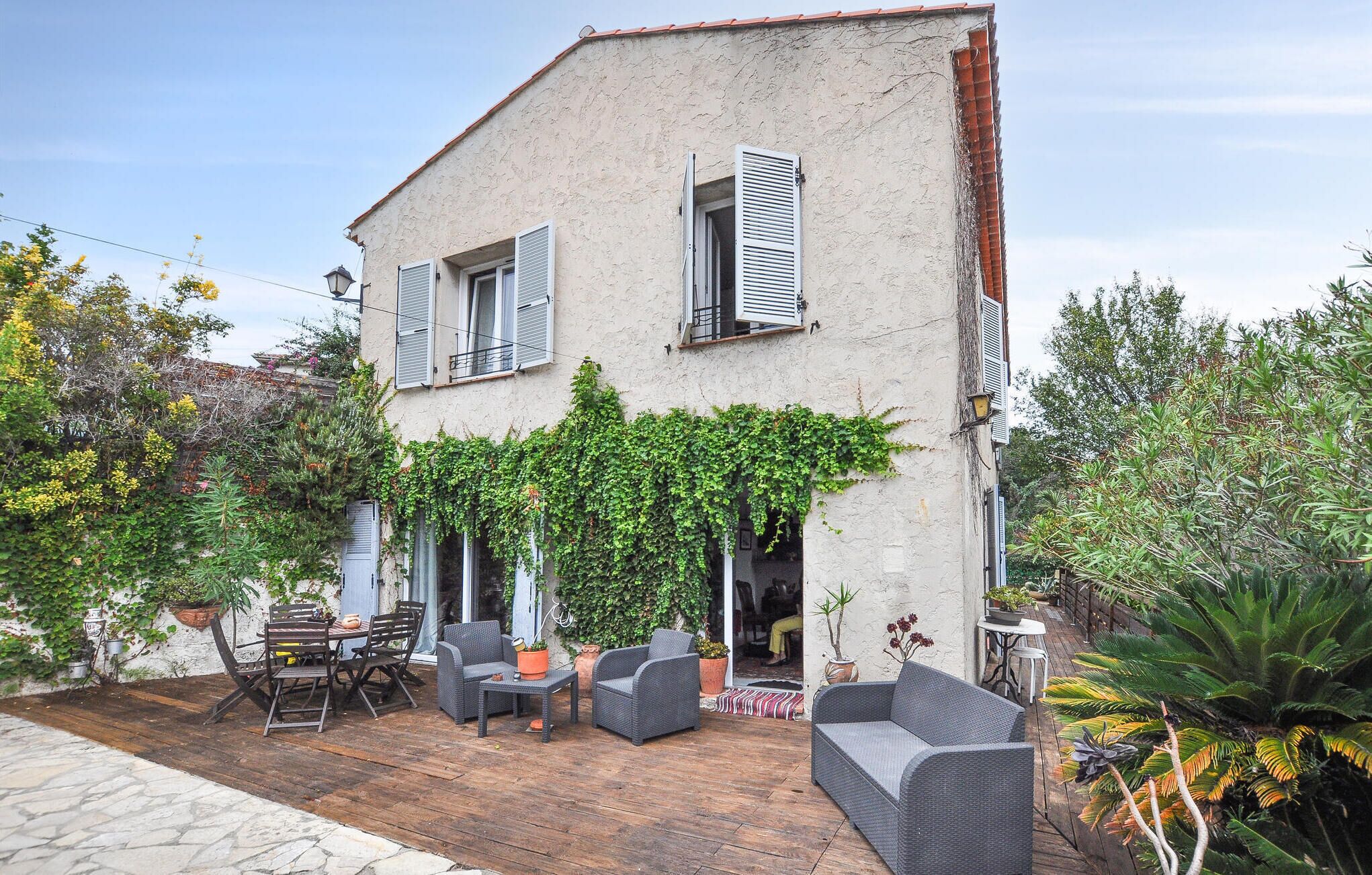 A two-story building with a stone facade, a balcony with wicker furniture, and a garden area with various plants.