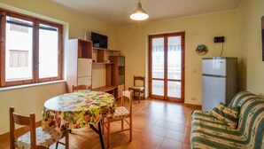 A dining area with a round table covered in a fruit-patterned tablecloth, surrounded by wooden chairs. There is a wooden cabinet with shelves, a television on top, and a refrigerator to the right. - Stunning apartment in Nocera Terinese (Nocera Terinese)