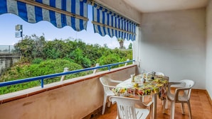 A balcony with a table set for dining, white plastic chairs, a striped awning, and a view of greenery and a bridge. - Stunning apartment in Nocera Terinese (Nocera Terinese)