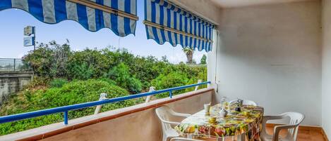 A balcony with a table set for dining, white plastic chairs, a striped awning, and a view of greenery and a bridge.